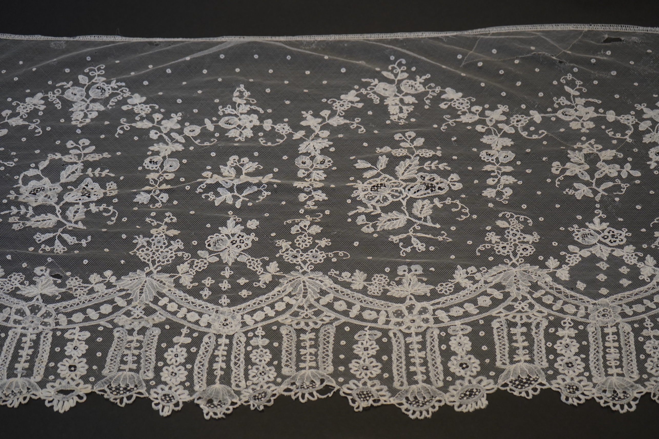 A deep elaborately designed flounce of mid 19th century Brussels bobbin appliquéd lace on net, with finished lace ends, worked with an intricate floral border below large floral cartouches framed in similar motifs and al